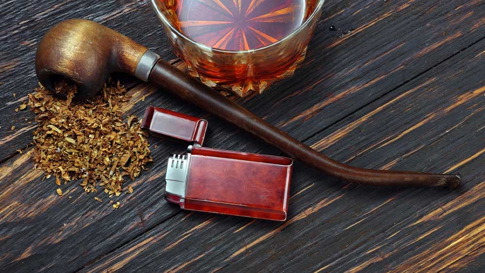 Pipa de fumat tutun impreuna cu o bricheta speciala de aprins pipa pe o masa cu tutun si un pahar de bautura